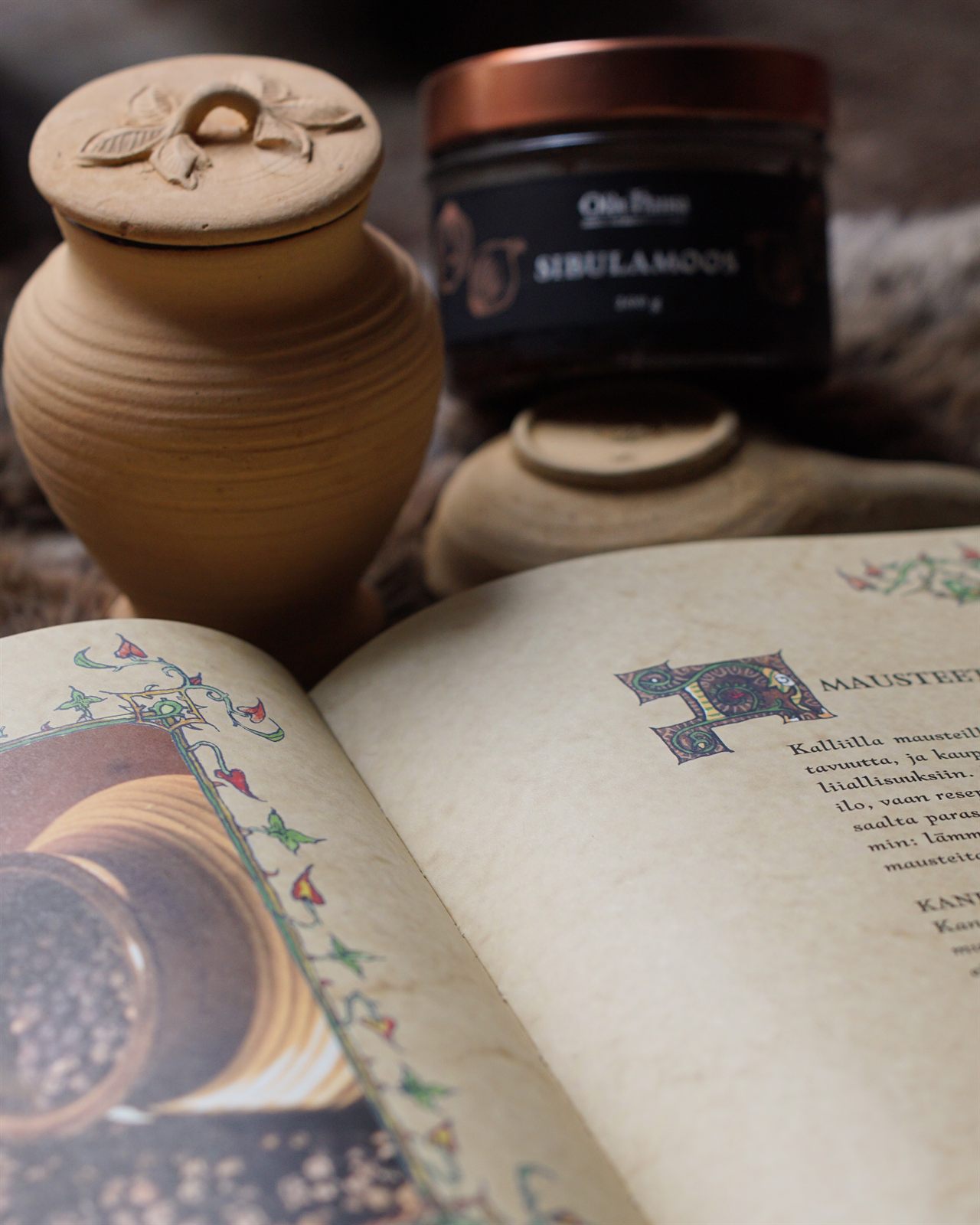 A lidded clay pot and a jar of Olde Hansa spice beside an open illuminated book